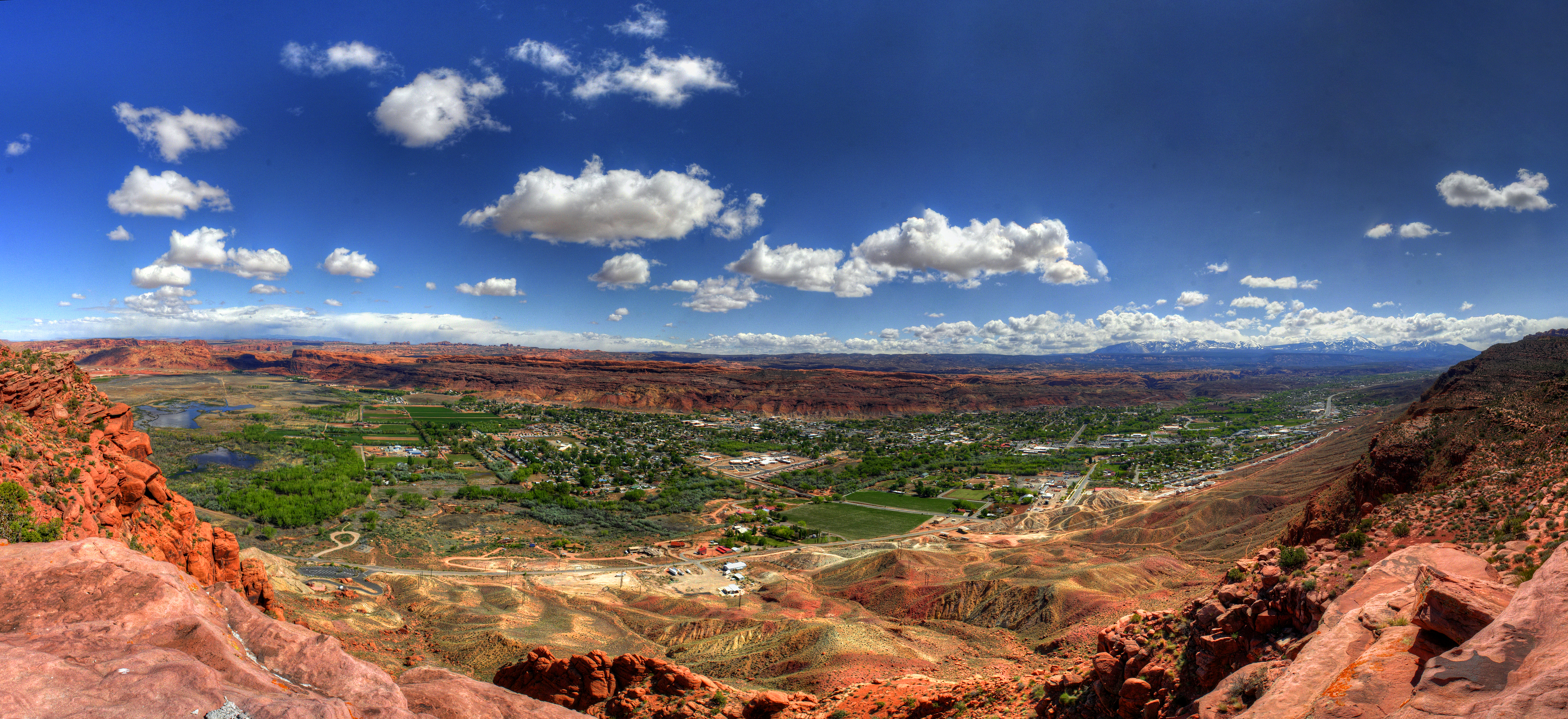 Panoramic view of the town of Moab. Bright blue sky, deep orange cliffs, and the green valley below.